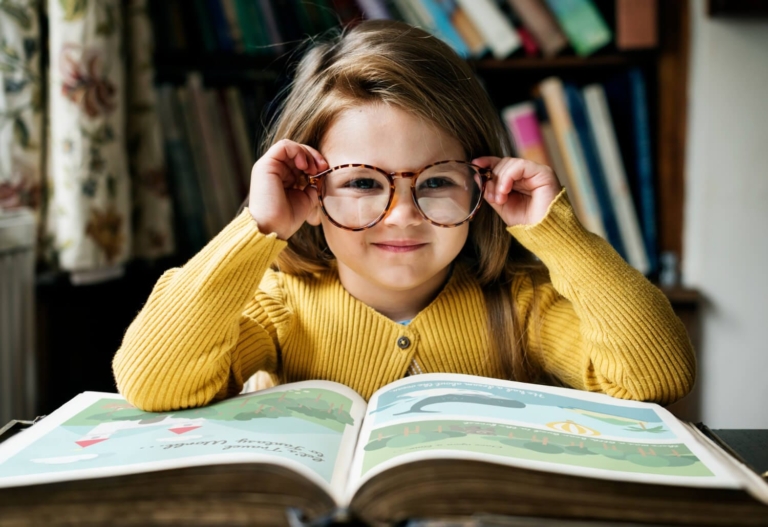 A young girl with light brown hair is adjusting her round, tortoiseshell glasses and looking directly at the camera, with an open book about web design storytelling in front of her, in a room with a bookshelf filled with books.