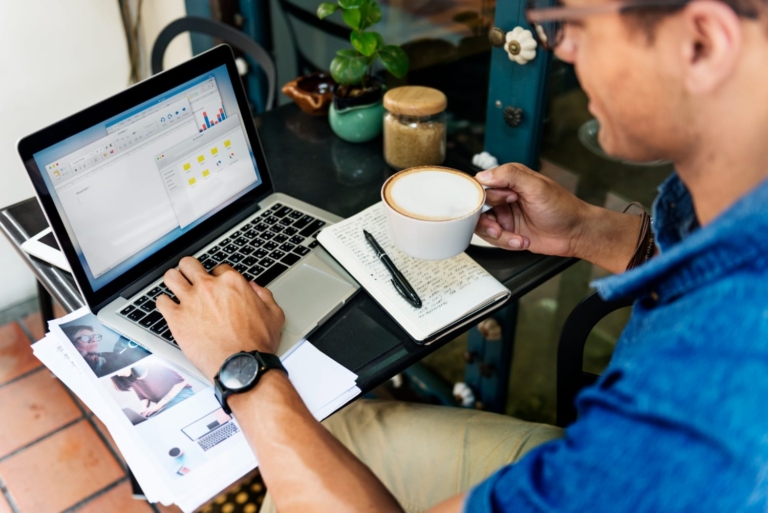 Man in blue shirt working on a laptop with analytics displayed on screen, holding a cup of coffee, with notes and printouts beside him.
